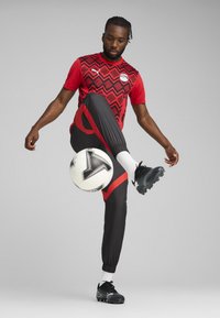 Rdeča in črna atletska majica s kratkimi rokavi in črnimi trenirko z rdečimi poudarki; igralec ravnotežno drži nogometno žogo.