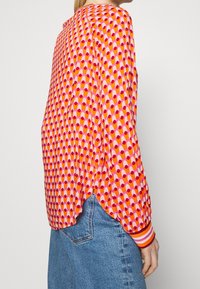 Nainen, joka on pukeutunut pitkähihaiseen paitaan, jossa on vaaleanpunaisia, oransseja ja punaisia geometrisiä kuvioita, yhdistettynä sinisiin farkkuihin, näkyy sivulta ja takaa.