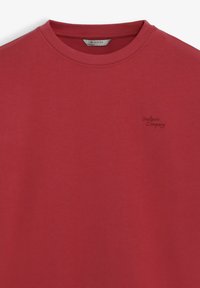T-shirt en coton rouge avec un col rond et des manches courtes. Présente un logo discret "Scalpers Company" brodé en rouge foncé.