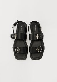 Svarte sandaler med to brede stropper med rektangulære spenner, flate såler og åpen design. Glatt lærtekstur, minimalistisk stil.