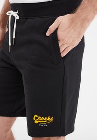 Pantalones cortos negros con cintura ajustable, con bolsillos laterales y un logo amarillo "Creeks" en la parte inferior izquierda. Textura de tela suave.