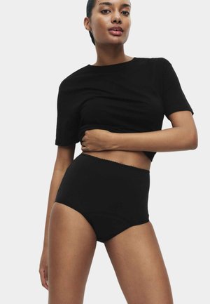 Frau trägt schwarzes bauchfreies T-Shirt und hoch taillierte schwarze Unterwäsche, steht vor einem einfarbigen hellen Hintergrund mit einem Arm über der Taille.