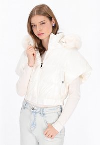 Biała kurtka puchowa o krótkim kroju, z kapturem wykończonym sztucznym futrem, przednim zamkiem błyskawicznym oraz krótkimi rękawami. Noszona na jasnej bluzce.
