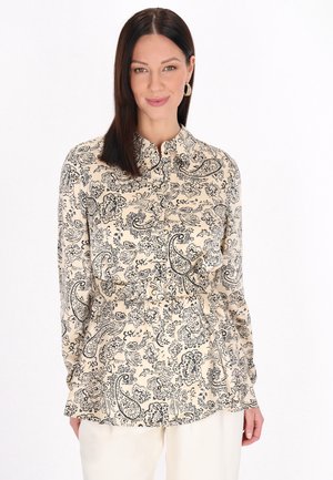Frau mit glatten dunklen Haaren trägt eine langärmelige beige Bluse mit schwarzen Blumen- und Paisley-Mustern, kombiniert mit hellen Hosen.