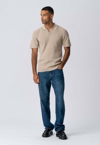 Beige kortærmet polo shirt med V-udskæring, parret med blå denimjeans og sorte plateau sko. Enkel, ribbet tekstur.