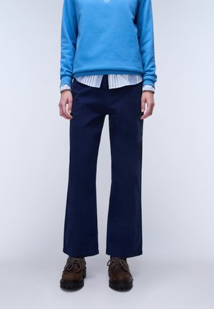 Person trägt dunkelblaue Hose mit geradem Bein, braune Schnürschuhe mit dicken Sohlen, ein hellblau gestreiftes Hemd und einen blauen Sweatshirt.