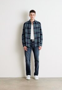 Camisa de franela a cuadros en azul marino y verde sobre una camiseta sin mangas blanca, combinada con jeans de mezclilla oscuros y zapatillas blancas. Diseño simple, ajuste casual.
