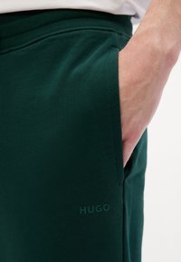 Pantaloni della tuta verdi con una consistenza morbida. Presentano tasche laterali e un logo "HUGO" tono su tono sulla gamba sinistra. Vita elasticizzata per una vestibilità ottimale.