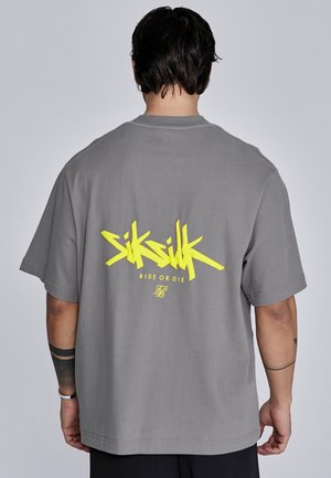 Camiseta gris de gran tamaño con un llamativo texto en estilo graffiti amarillo "SikSilk" y "Ride or Die" en la parte posterior. Diseño de mangas cortas y cuello redondo.