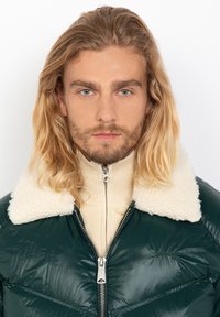 Green puffer jacket with a cream shearling collar, featuring a front zipper and subtle stitching details. Worn over a light beige turtleneck.
