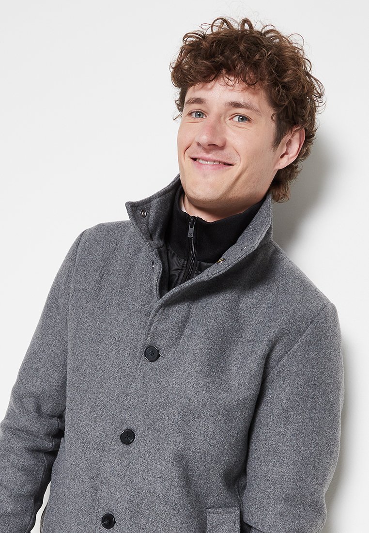 Szary płaszcz z mieszanki wełny z wysokim kołnierzem, zapięciem na guziki i kieszeniami bocznymi. Tkanina ma gładką strukturę i wyraźny kształt.