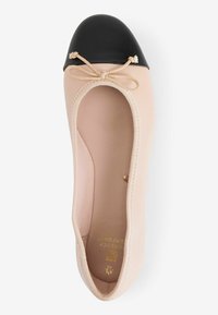 Beige ballet flats with a black leather cap toe, bow detail, and soft fabric lining. They feature a low-profile design and a rounded shape.