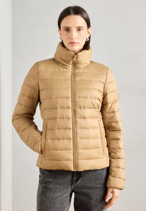 Beige gewatteerde jas met een hoge kraag, horizontaal stikselpatroon, volledige rits en zijzakken. Draagt bij grijze spijkerbroek tegen een neutrale achtergrond.