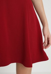 Vestido rojo en línea A, hecho de una tela suave, con un dobladillo de falda acampanada y detalles de costura visibles. Mano reposando en el borde de la falda.