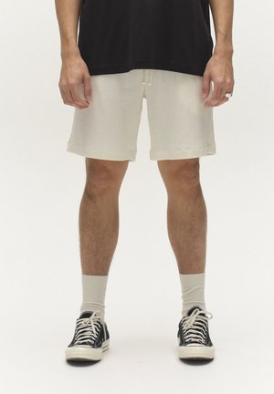 Persoon die crèmekleurige korte broeken met een trekkoord, zwarte sneakers, crèmekleurige sokken en een zwart shirt draagt, staand op een effen witte achtergrond.