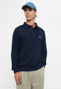 Mann trägt ein dunkelblaues langärmliges Poloshirt mit grünem Krokodil-Logo, beige Hose und eine hellblaue Baseballmütze, schaut zur Seite.