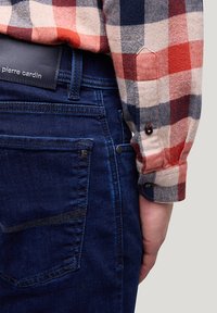 Ärmel eines karierten Hemdes in Rot, Beige und Marineblau liegt neben einer dunkelblauen Jeans aus Denim mit einem schwarzen Lederetikett von Pierre Cardin am Bund.