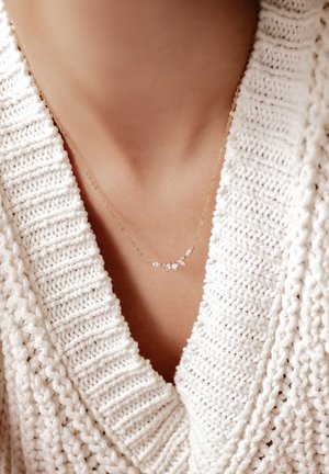 Zarte Goldkette mit blattförmigen klaren Edelsteinen. Getragen zu einem cremfarbenen, gestrickten Pullover mit strukturiertem, offenem Strickmuster.