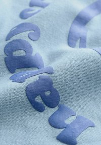 Produit textile en bleu clair avec des lettres ludiques en bleu foncé. Le tissu a une surface lisse et légèrement texturée, créant un contraste entre les couleurs.