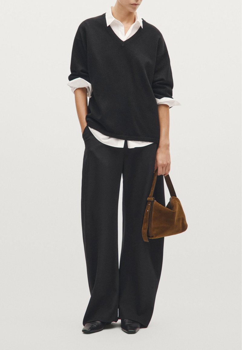 Sort oversized sweater med V-udskæring, hvid skjorte krave under, brede sorte bukser og en lille brun ruskinds håndtaske. Sorte spidse flade sko.