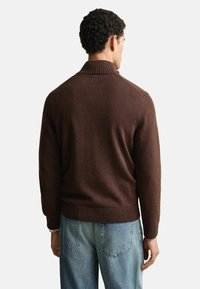 Brauner Strickpullover mit hohem Kragen, geripptem Saum und Ärmeln, kombiniert mit hellblauen Jeans. Der Stoff wirkt weich und strukturiert.