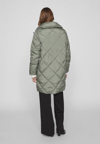 Grüner gepolsterter Mantel mit Rautenmuster, weit geschnittenem Kragen und langen Ärmeln. Über schwarzen Hosen getragen. Glatte, glänzende Stoffoberfläche.