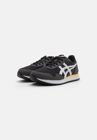 Black athletic shoes made of mesh and synthetic materials. Features white stripes on the sides, textured black laces, and a yellow accent on the sole.