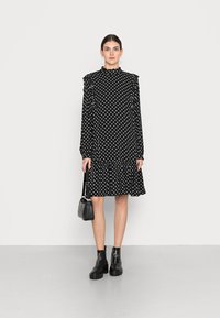 Vestido preto de bolinhas com ombros franzidos, gola alta e bainha rodada. Combinado com botas ankle pretas e uma pequena mala preta.