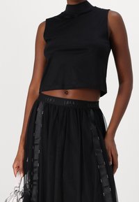 Femme portant un crop top noir sans manches et une jupe en tulle noir avec une ceinture de marque, tenant un petit sac à main en argent.