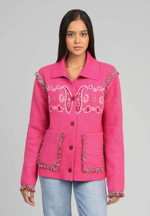 Femme portant une veste boutonnée rose vif avec des broderies blanches et des détails à franges, associée à un jean bleu clair, debout devant un fond gris.
