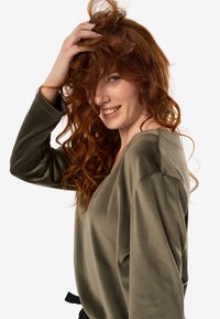 Luźna bluzka w oliwkowej zieleni z długimi rękawami i głębokim dekoltem w kształcie litery V. Gładki materiał z lekkim połyskiem, ozdobiona czarnym paskiem wiązanym w talii.