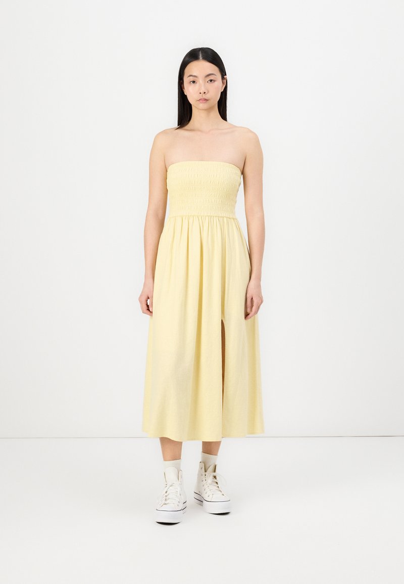 Rochie galbenă fără bretele, cu un corset pliat, fustă fluidă, fante pe lateral și asortată cu adidași albi. Textură netedă și design până la genunchi.