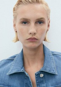 Lys blond hår med en tekstureret, lagdelt stil; iført en blå denimjakke med knap og krave; hud med lyse fregner; neutral baggrund.
