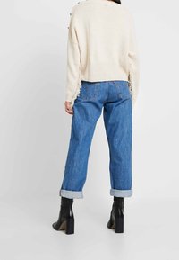 Beżowy sweter dziany z frędzlami, w zestawie z niebieskimi dżinsami z podwiniętymi nogawkami i czarnymi botkami na blokowym obcasie.