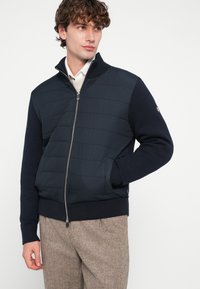 Navy zila hibrīda jaka ar polsterētu priekšējo daļu, ribotu aproču un apkakles malu, rāvējslēdža aizdare un pieguļošs dizains. Valda valkāta ar bēša rakstainām biksēm.