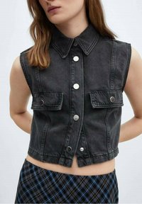 Schwarze Denim-Weste ohne Ärmel mit klassischem Kragen, silbernen Druckknöpfen, zwei vorderen Taschen und einem cropped Design. Darunter ein karierter Rock.