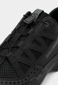 Μαύρο αθλητικό παπούτσι με άνω μέρος από δίχτυ, με λαστιχένιες λεπτομέρειες, εφαρμοστό σύστημα κορδονιών και υφασμένο πέλμα για κράτημα και υποστήριξη.