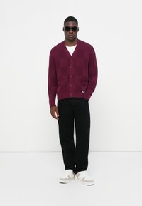 Cardigan lavorato a maglia bordeaux con motivo a quadri, chiusura con bottoni e due tasche frontali, abbinato a pantaloni neri e sneakers bianche.