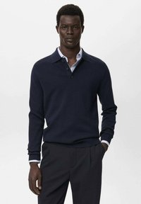 Man in een marineblauwe langemouwen polo over een lichtblauw overhemd met kraag en donkere broeken, staande met één hand in de broekzak.
