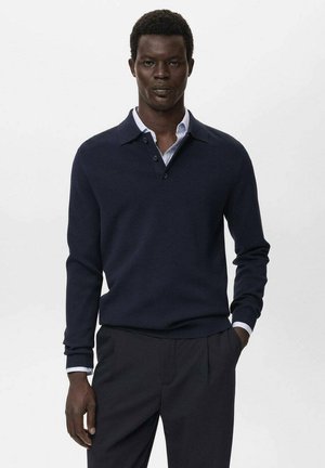 Man in een marineblauwe langemouwen polo over een lichtblauw overhemd met kraag en donkere broeken, staande met één hand in de broekzak.