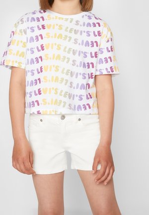 Enfant portant un t-shirt court blanc avec un texte multicolore répété "Levi's" et un short en denim blanc, debout devant un fond uni.