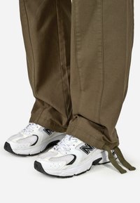 Olive žalio atspalvio kelnės su tiesiu kirpimu, derinamos su baltais New Balance sportbačiais, turinčiais tinklinio akcentus ir juodus detalizavimus.