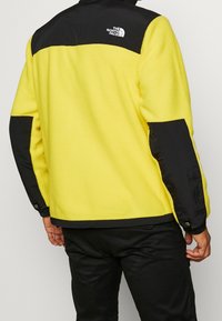 Muž v žlutočerné fleecové bundě North Face, zobrazený ze zadu, s černými kalhotami na jednobarevném pozadí.