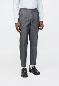 Calças cinzentas às riscas com um corte ajustado, apresentando um cós plano, passadores para cinto e uma vinca bem marcada. Usadas com sapatos pretos e meias claras.