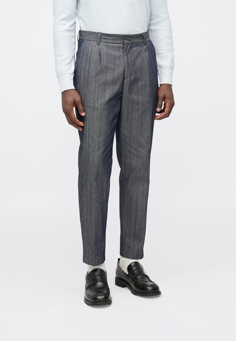 Calças cinzentas às riscas com um corte ajustado, apresentando um cós plano, passadores para cinto e uma vinca bem marcada. Usadas com sapatos pretos e meias claras.