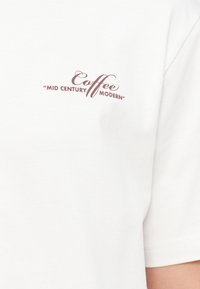 Λευκό T-shirt με καφέ κείμενο στο στήθος που αναγράφει: "Coffee 'Mid Century Modern'" σε μικτή γραφή και κεφαλαία γράμματα.