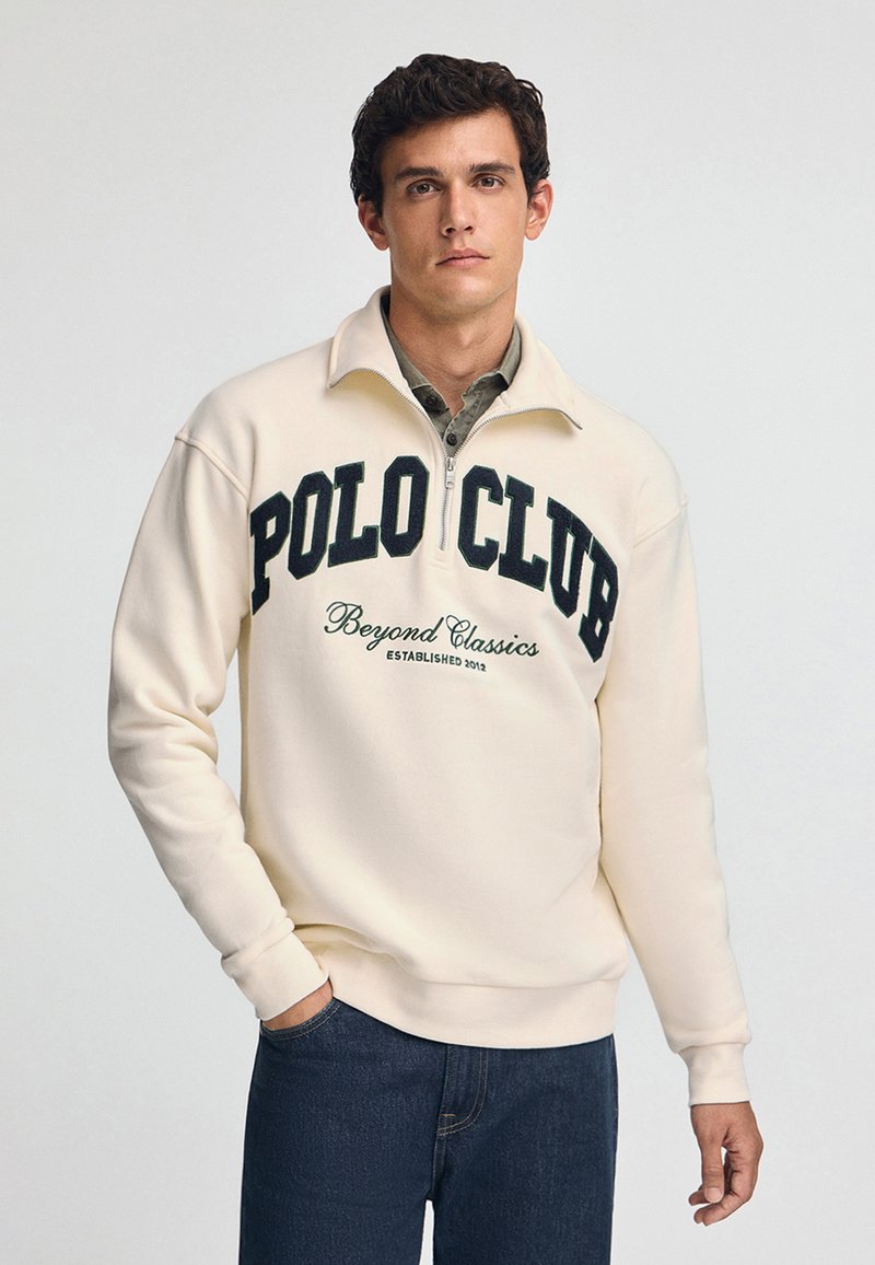Krämfärgad zip-up sweatshirt med "POLO CLUB"-logotyp i mörkgrön. Ribbad muddar och midja, bärs med mörka jeans. Slät textur.