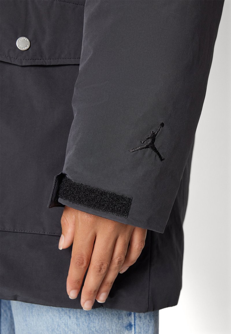 Vêtements d'extérieur noirs avec un poignet en velcro, mettant en avant un logo Jumpman brodé sur la manche. Le tissu semble lisse et légèrement texturé.