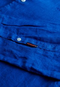 Camisa de lino azul con botones blancos, una pequeña etiqueta "Superdry" en negro y naranja, y un tejido texturizado con sutiles arrugas.