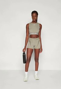 Olívazöld Nike Pro sportfelső és rövidnadrág, márkás elasztikus pánttal. A modell egy fekete vízpalackot tart, és világos sportcipőt visel.